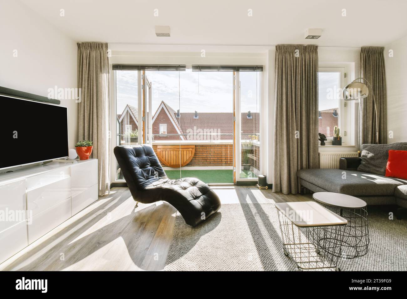 Schwarzer Ledersessel mit Sofa und Couchtischen vor dem Fernseher durch großes helles Fenster im modernen Wohnzimmer Stockfoto