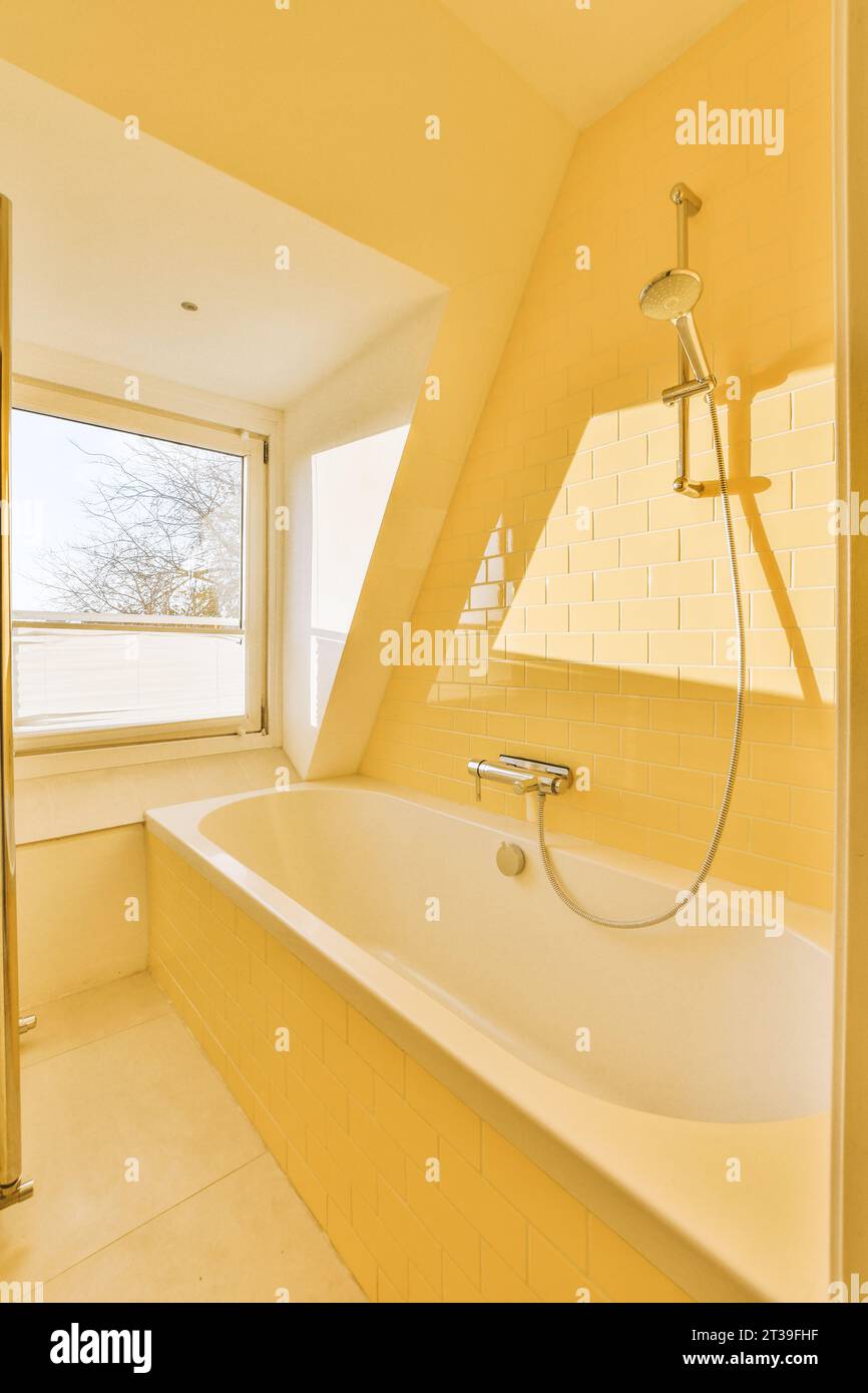 Weiße Badewanne mit Edelstahlhahn am Fenster im Badezimmer mit gelben Fliesenwänden in einem modernen Apartment Stockfoto