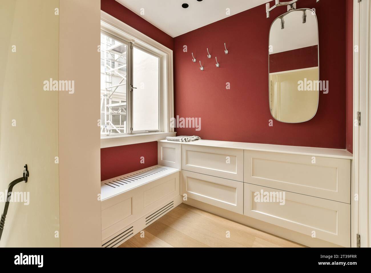 Spiegel und Haken an der roten Wand über weißen Schränken am Fenster in einem kleinen Zimmer in einem modernen Apartment Stockfoto