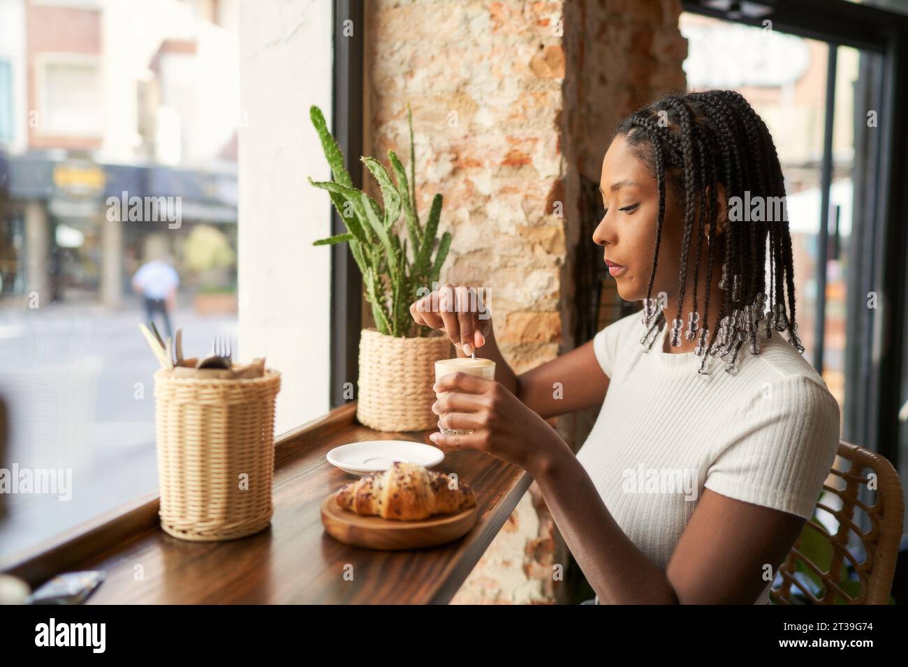 Seitenansicht einer jungen, nachdenklichen afroamerikanischen Frau, die nach unten blickt, während sie am Schreibtisch in der Kantine mit Croissant auf einem Teller saß und heißes Getränk im Glas verkostete Stockfoto