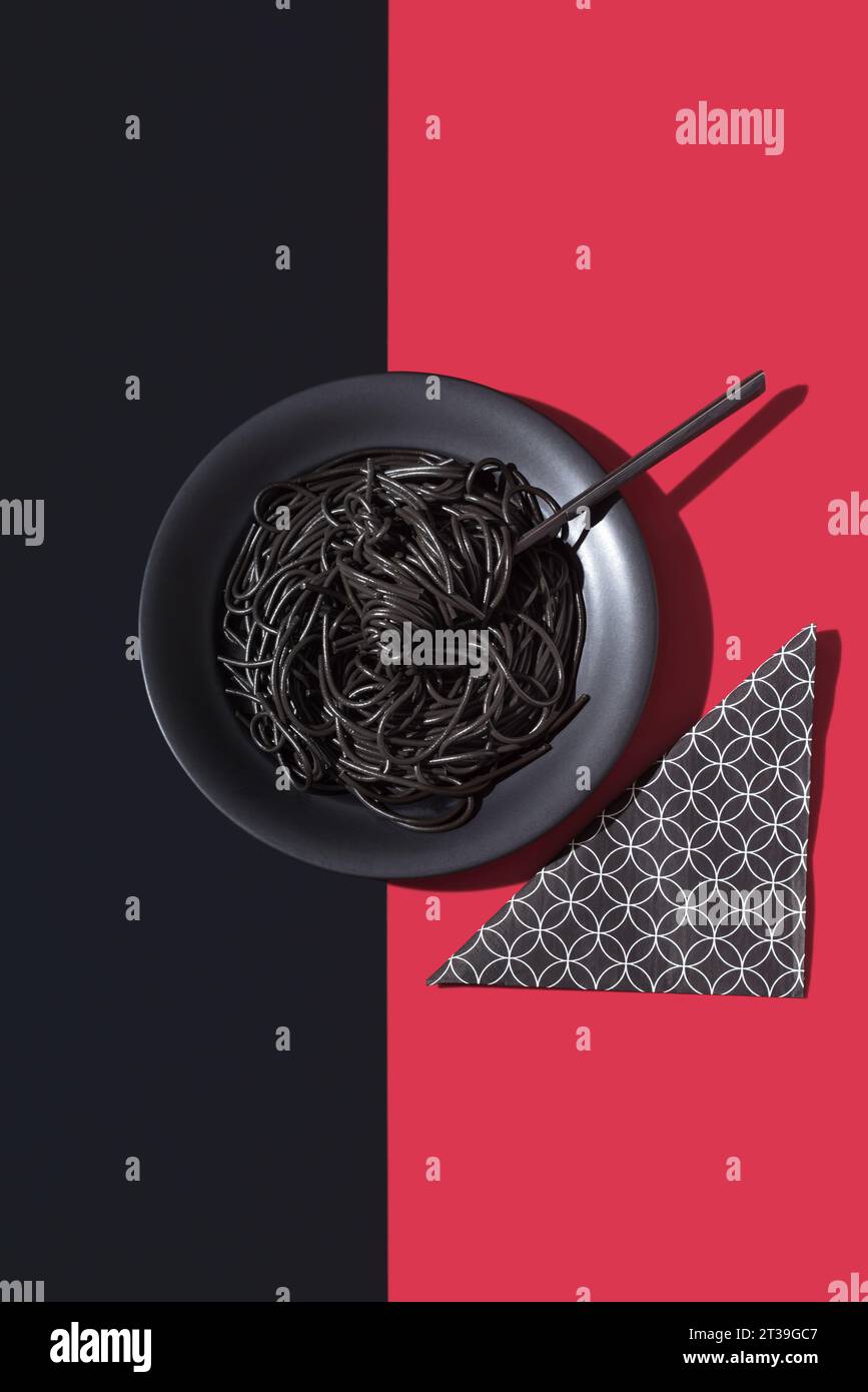 Draufsicht auf schwarze Spaghetti in einer Schüssel mit Gabel auf schwarzem und rotem Hintergrund in der Nähe der Serviette Stockfoto