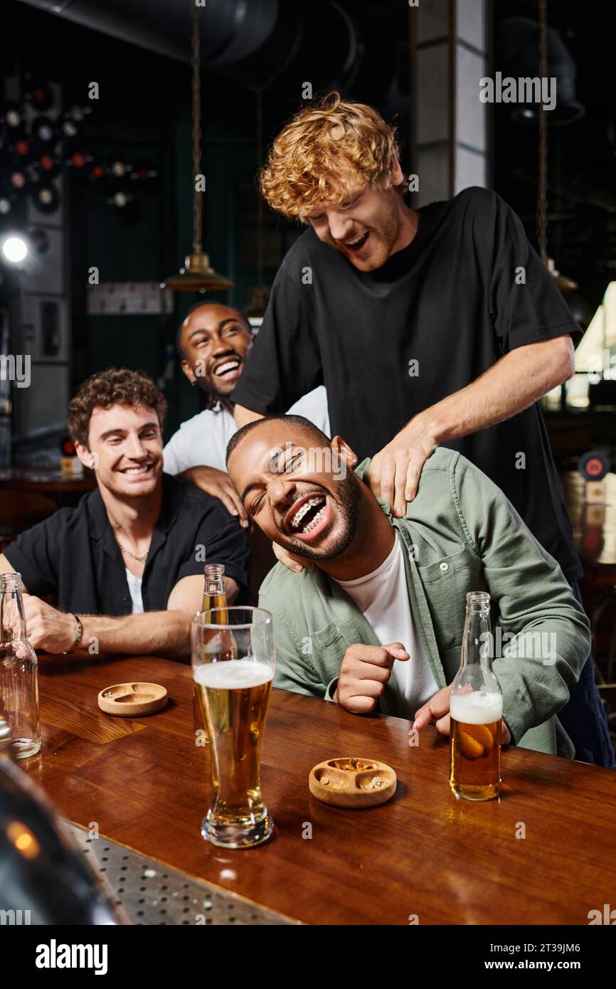 Rothaariger Mann, der glückliche afroamerikanische Freund während der Junggesellenabschied in der Bar kitzelt, männliche Freundschaft Stockfoto