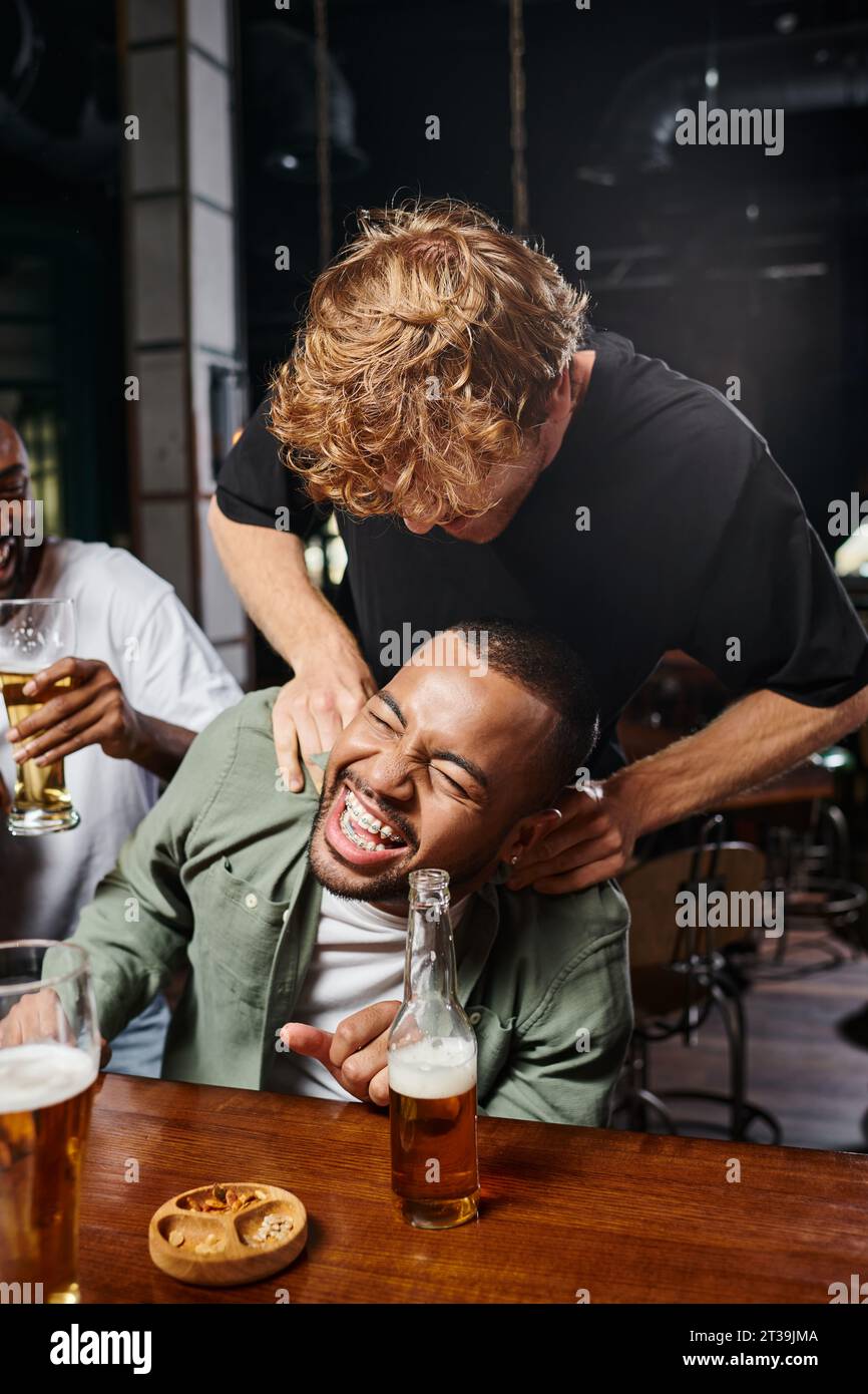 Rothaariger Mann, der fröhlich afroamerikanischen Bräutigam während der Junggesellenabschied in der Bar kitzelt, männliche Freundschaft Stockfoto
