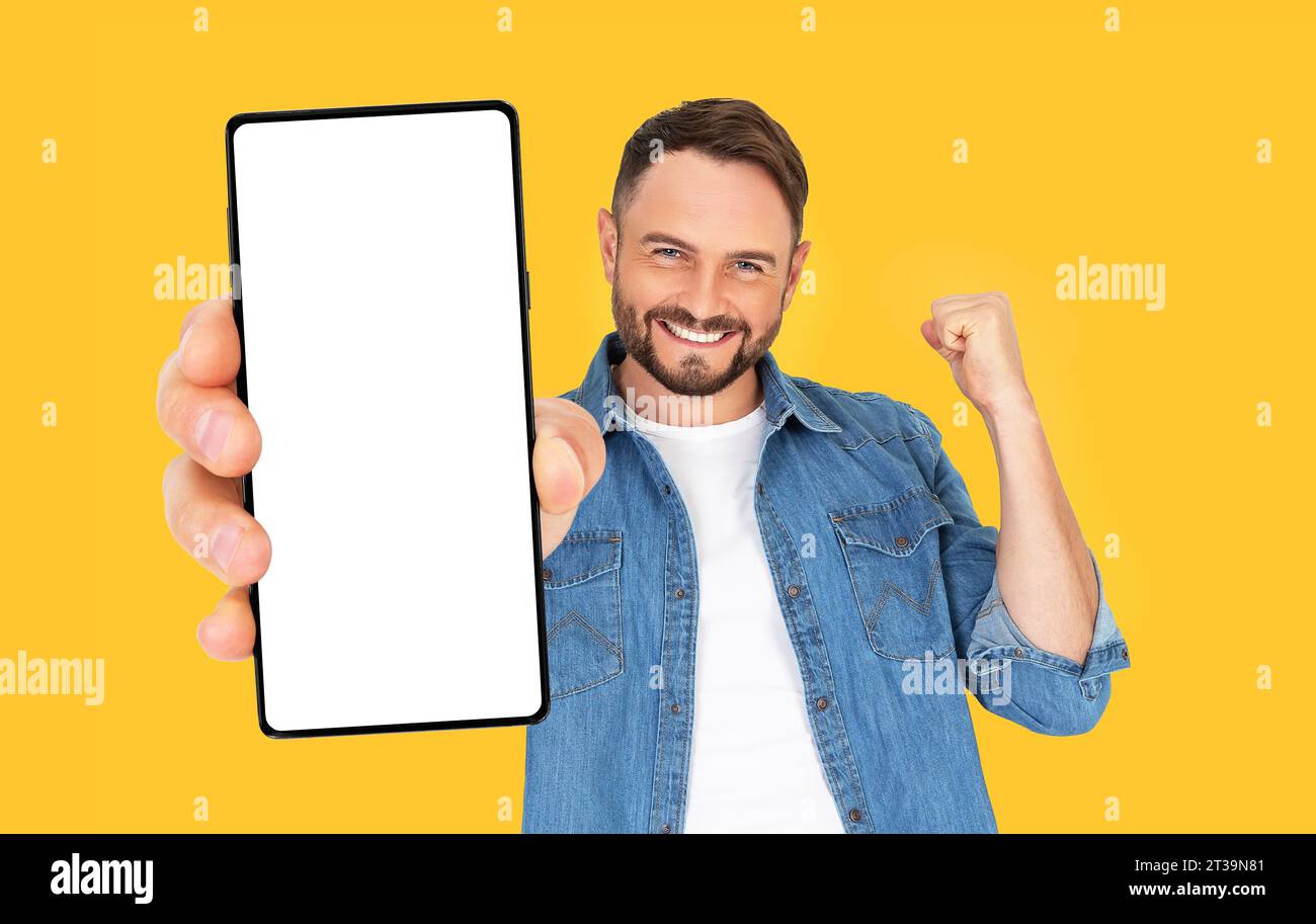 Hübscher junger Mann, der ein Smartphone-Modell mit einem leeren Bildschirm hält und auf einem orangefarbenen Hintergrund lächelt. Erfolg feiern. Gadget mit Leerzeichen f Stockfoto