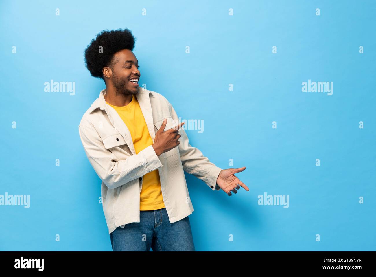 Überraschter afro-amerikanischer Mann lächelt mit Händen, die auf den Raum zeigen, in hellblauem Farbstudio fotografiert isolierter Hintergrund Stockfoto