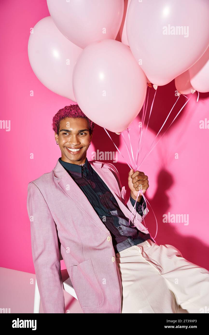 Gut aussehender Mann mit gelockten rosa Haaren, der auf weißem Stuhl posiert, mit Ballons in der Hand, Puppe wie Stockfoto