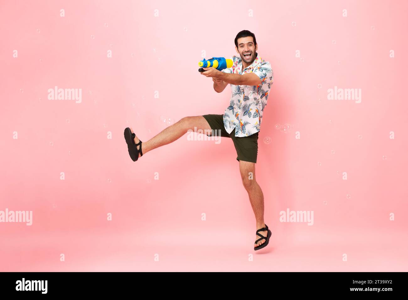 Hübscher, lächelnder junger kaukasischer Touristenmann, der mit einer Wasserpistole spielt und im rosafarbenen Studio für das Songkran Festival in Thailand und im Süden springt Stockfoto