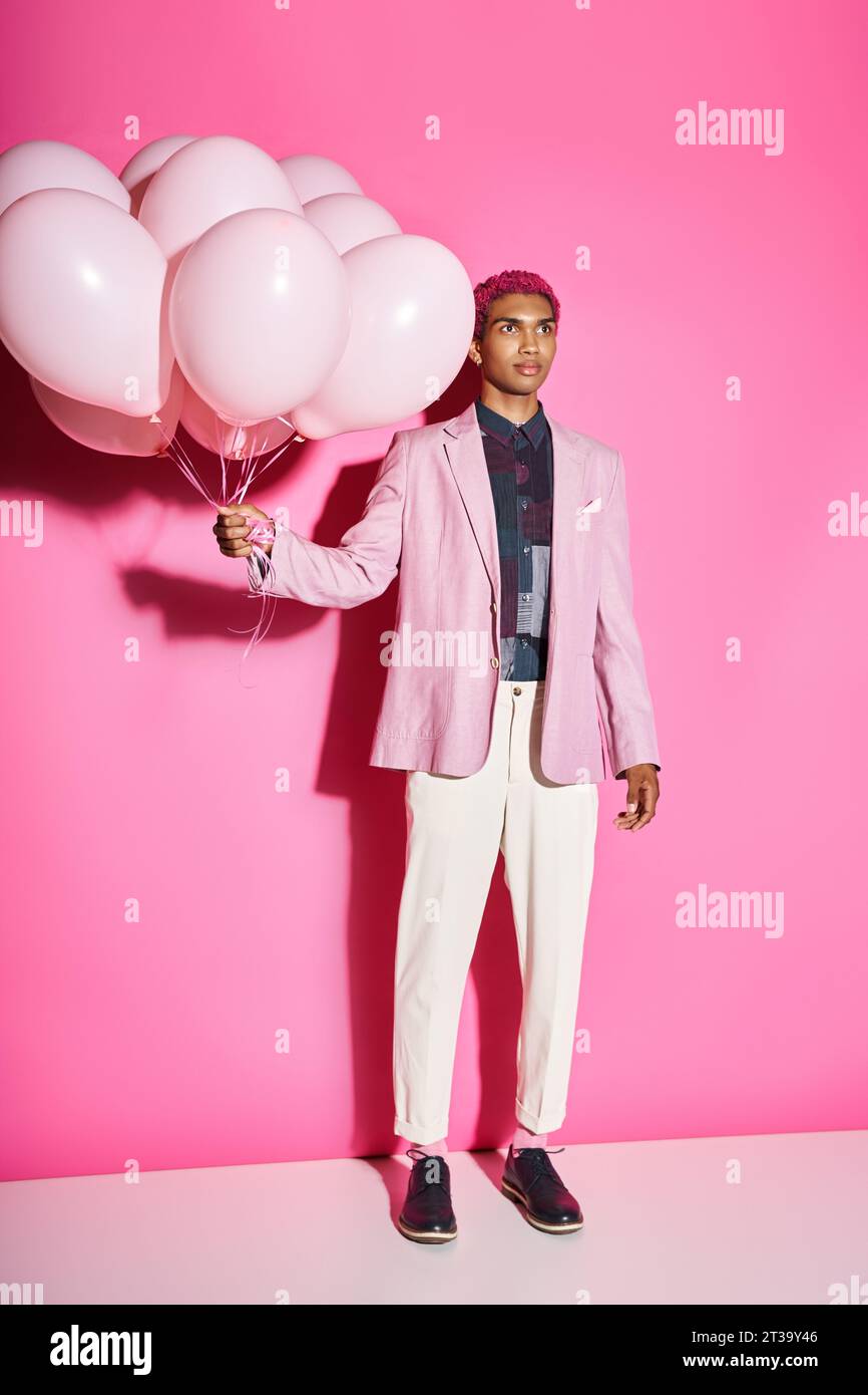 Hübscher Mann mit lockigen rosa Haaren in rosa Blazer posiert mit Ballons auf rosa Hintergrund, Puppe wie Stockfoto