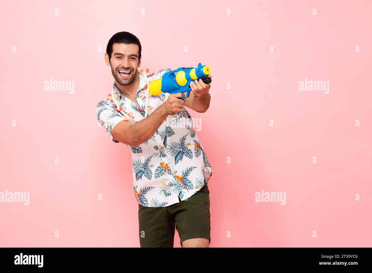 Fröhlich lächelnder junger kaukasischer Touristenmann, der mit Wasserpistole im rosa Studiohintergrund für das Songkran Festival Konzept in Thailand und Südosten spielt Stockfoto
