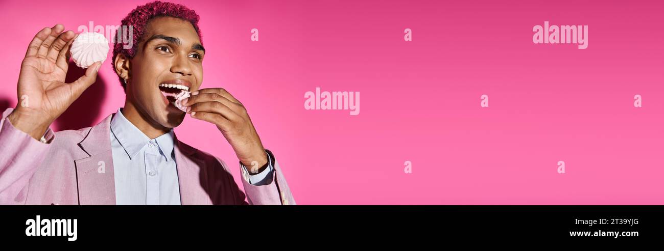 Hübsches, junges männliches Model mit lockigen rosa Haaren, das köstliche zefir isst und wegblickt, Banner Stockfoto
