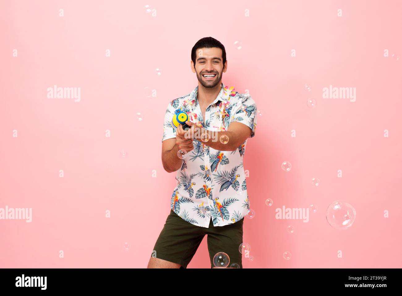 Fröhlich lächelnder junger kaukasischer Touristenmann, der mit Wasserpistole im rosa Studiohintergrund für das Songkran Festival Konzept in Thailand und Südosten spielt Stockfoto