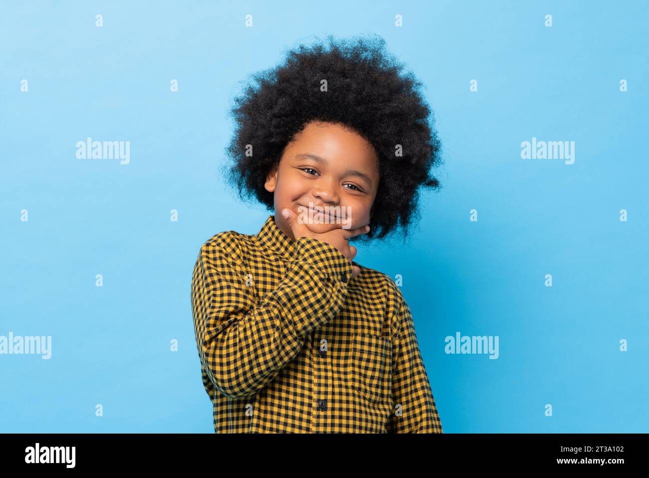 Niedlicher afrikanischer gemischter Afro-Junge posiert und lächelt mit Hand auf Kinn in blauer Farbe isolierter Hintergrund-Studio-Aufnahme Stockfoto