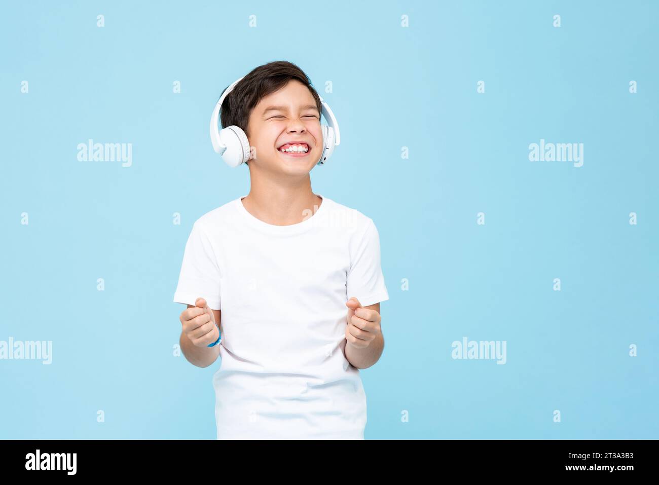 Glücklich lächelnde gemischte Rasse asiatischer Junge, der Kopfhörer trägt, hört Musik und klirrende Fäuste in isoliertem hellblauem Hintergrund Studioaufnahme Stockfoto