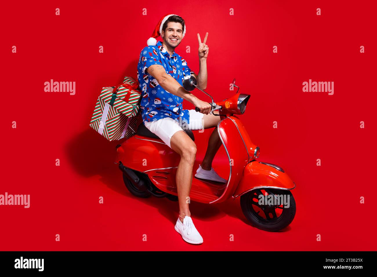 Foto in voller Länge von netten jungen männlichen präsentiert Lieferung Moped V-Zeichen tragen santa claus Druck x-MAS Kleid isoliert auf rotem Hintergrund Stockfoto