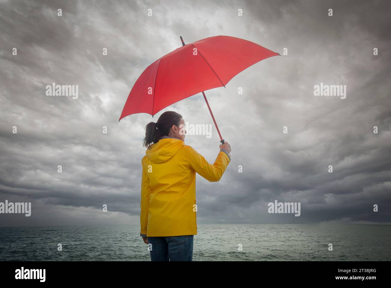 Eine Frau, die einen stürmischen Himmel sieht, einen roten Regenschirm hält und einen gelben Mantel trägt. Stürmisches Wetterkonzept. Stockfoto