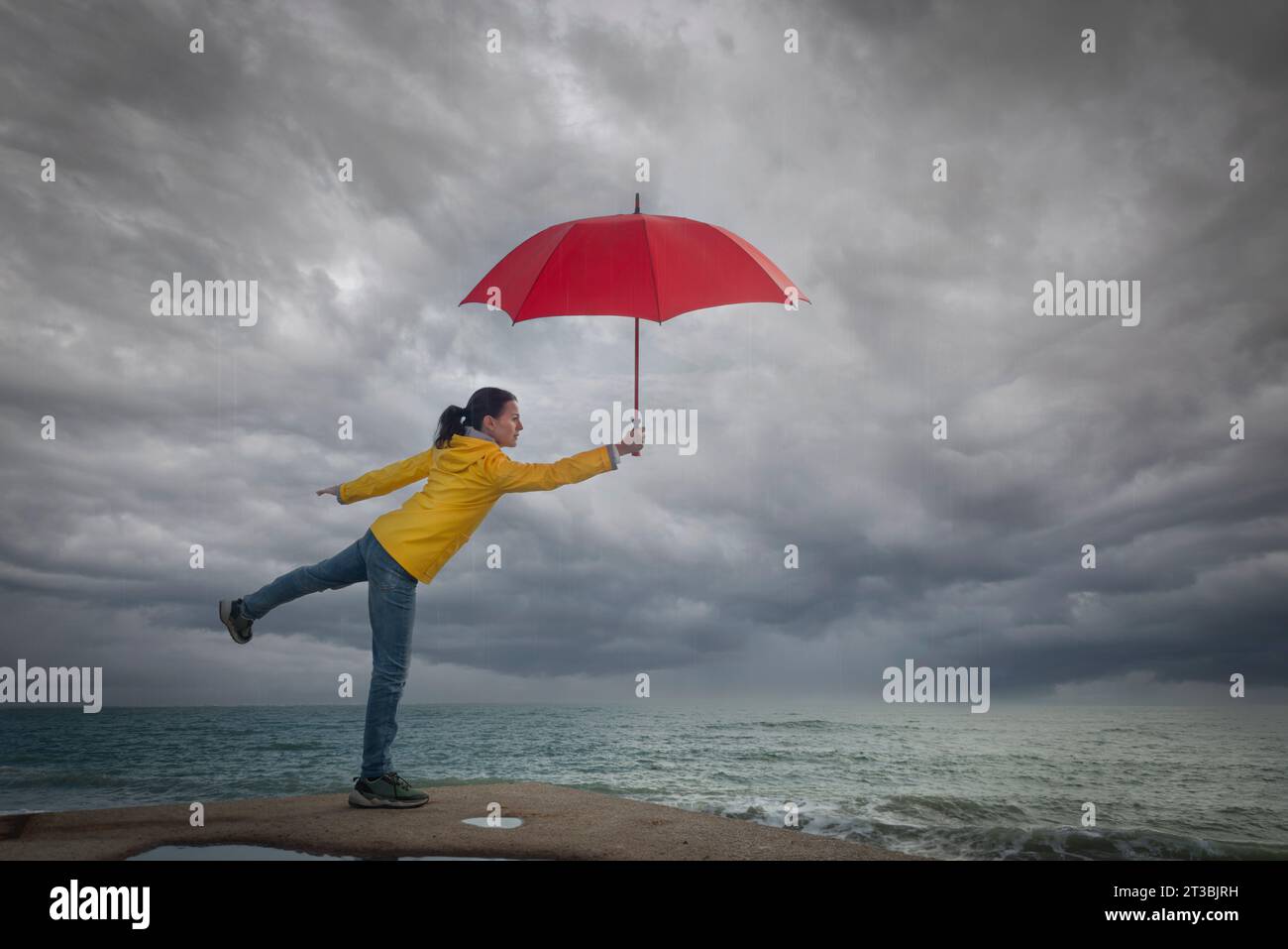 Eine Frau, die einen stürmischen Himmel sieht, einen roten Regenschirm hält und einen gelben Mantel trägt. Stürmisches Wetterkonzept. Stockfoto