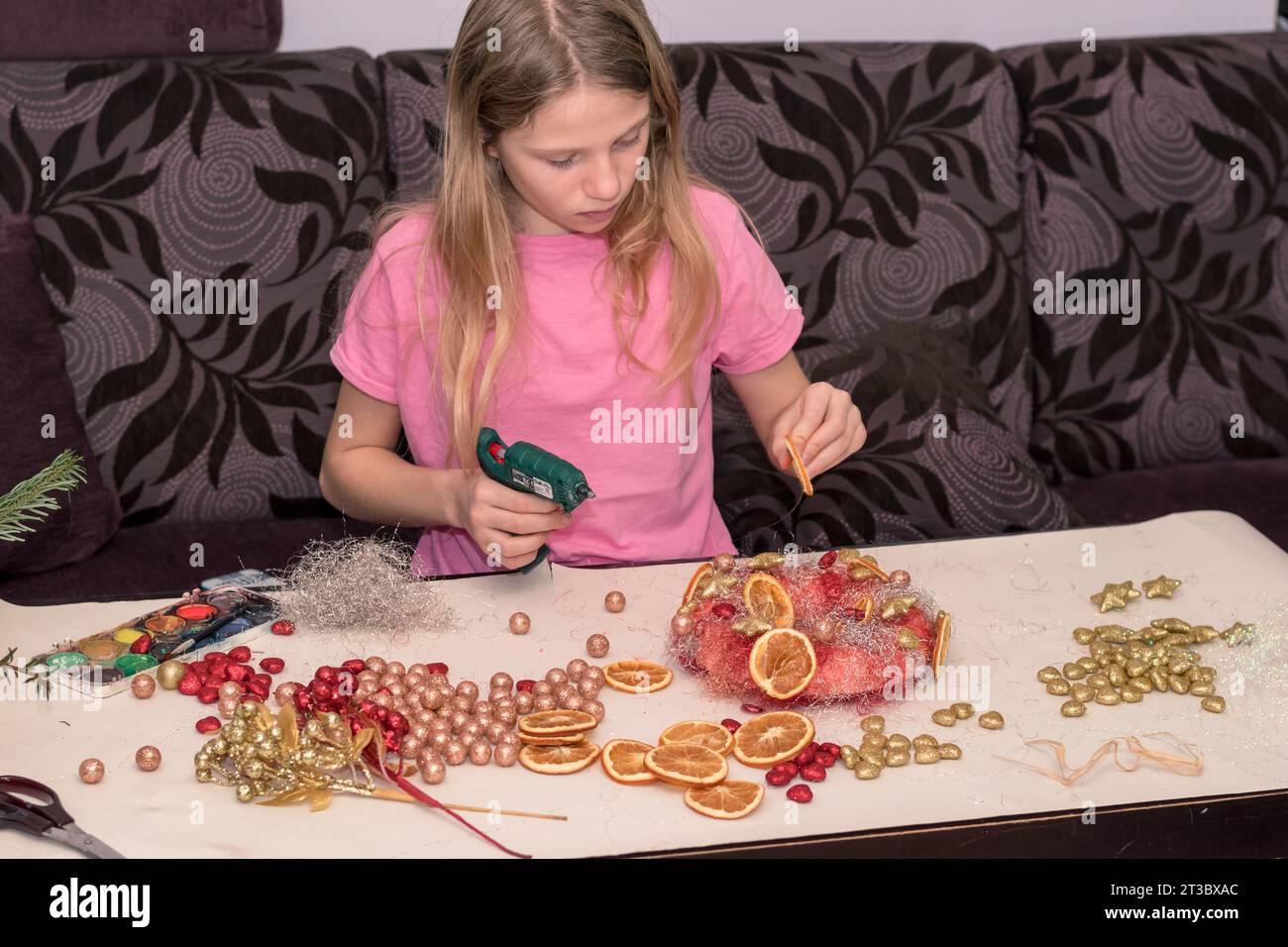 Niedliches Mädchen, das einen weihnachtskranz mit Kugeln und anderen bunten Glitzerdekorationen kreiert Stockfoto