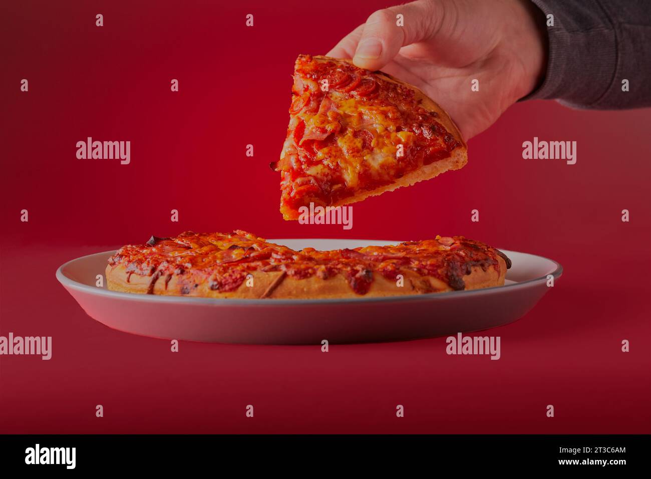 Frisch gebackene fleischige, käsige, tiefe Pfannenpizza, mit einem Stück, das mit rotem Hintergrund hochgehalten wird. Stockfoto