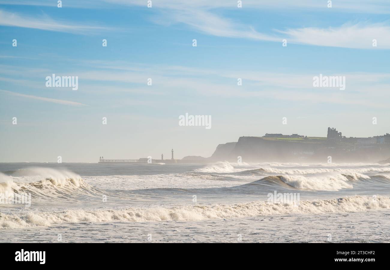 An einem frischen Herbstmorgen krachen große Wellen in die Nordseeküste zwischen Sandsend und Whitby, wobei die berühmte Abtei und der Pier am Horizont sichtbar sind. Stockfoto