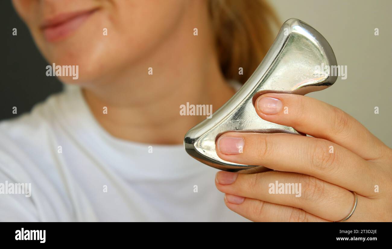 Junge kaukasische Frau lächelt und hält ein Metall-Gua-sie-Werkzeug, um das Gesicht zu heben Stockfoto