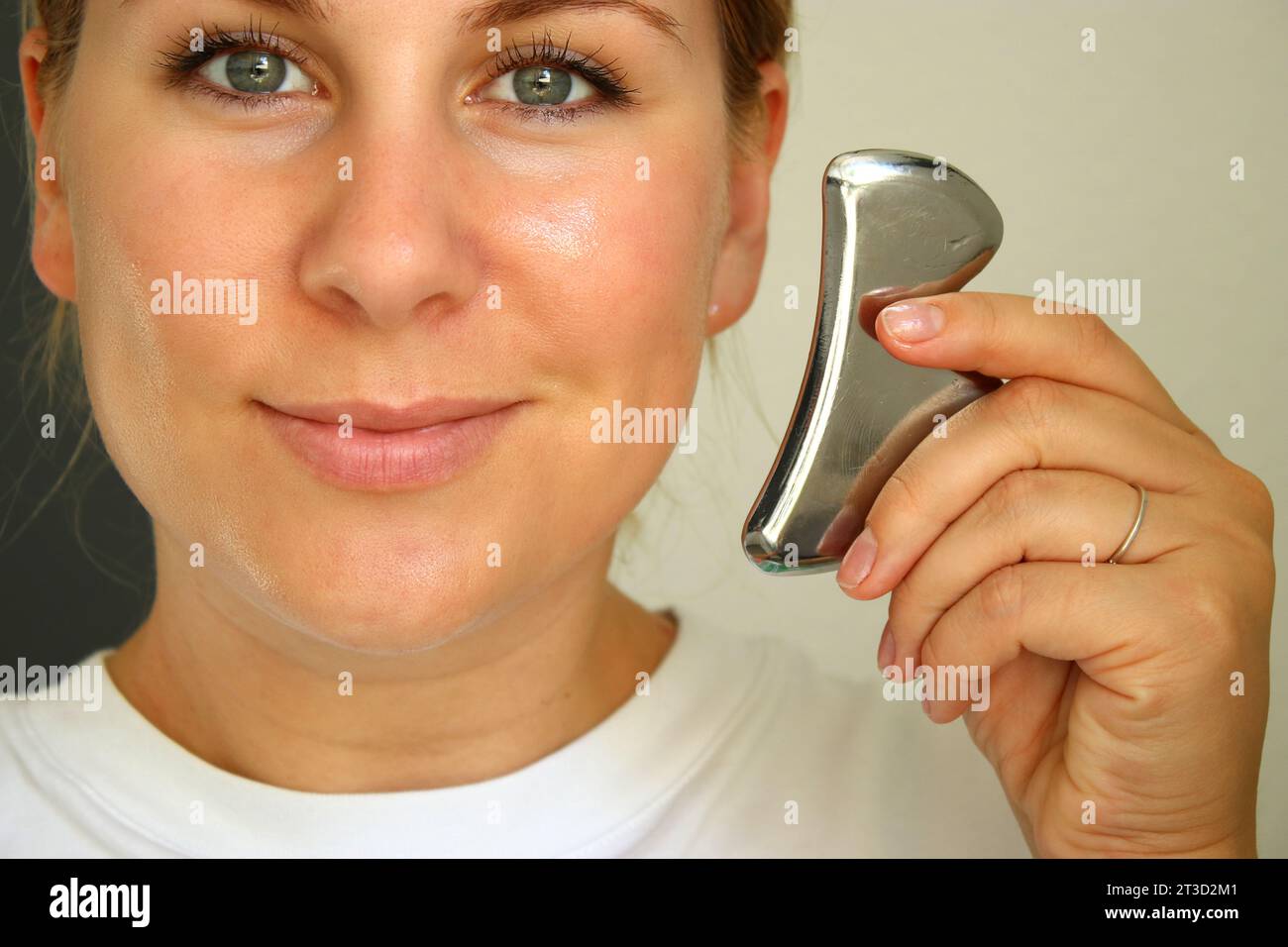 Schöne weiße Frau lächelt und hält ein Metall-Gua-sie-Werkzeug zum Anheben des Gesichts Stockfoto