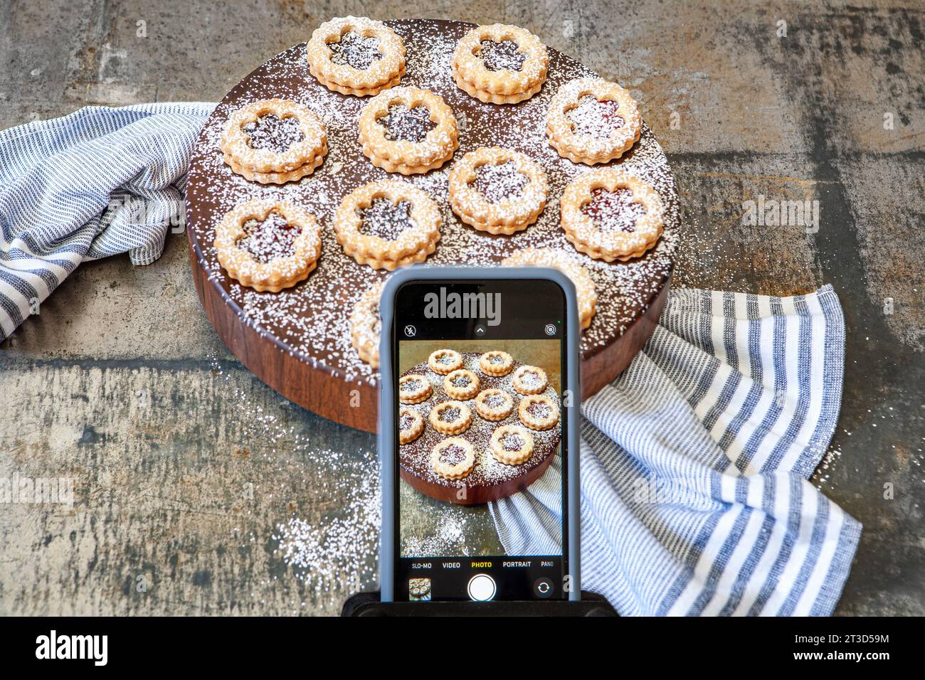 Hochwinkelansicht des iPhone, bei dem Linzer Torten mit Puderzucker auf rundem Holzbrett mit blauem Handtuch fotografiert werden Stockfoto