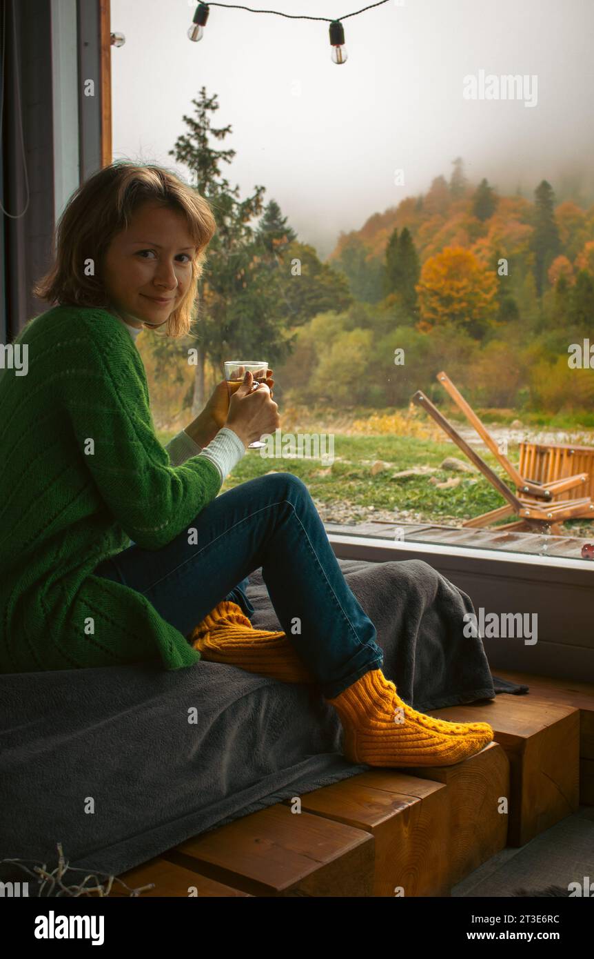 Mädchen auf einem Bett mit einer Tasse Tee hinter einem großen Fenster mit atemberaubender Herbstlandschaft. Frau, die draußen im Nebel in die Kamera blickt. Stockfoto