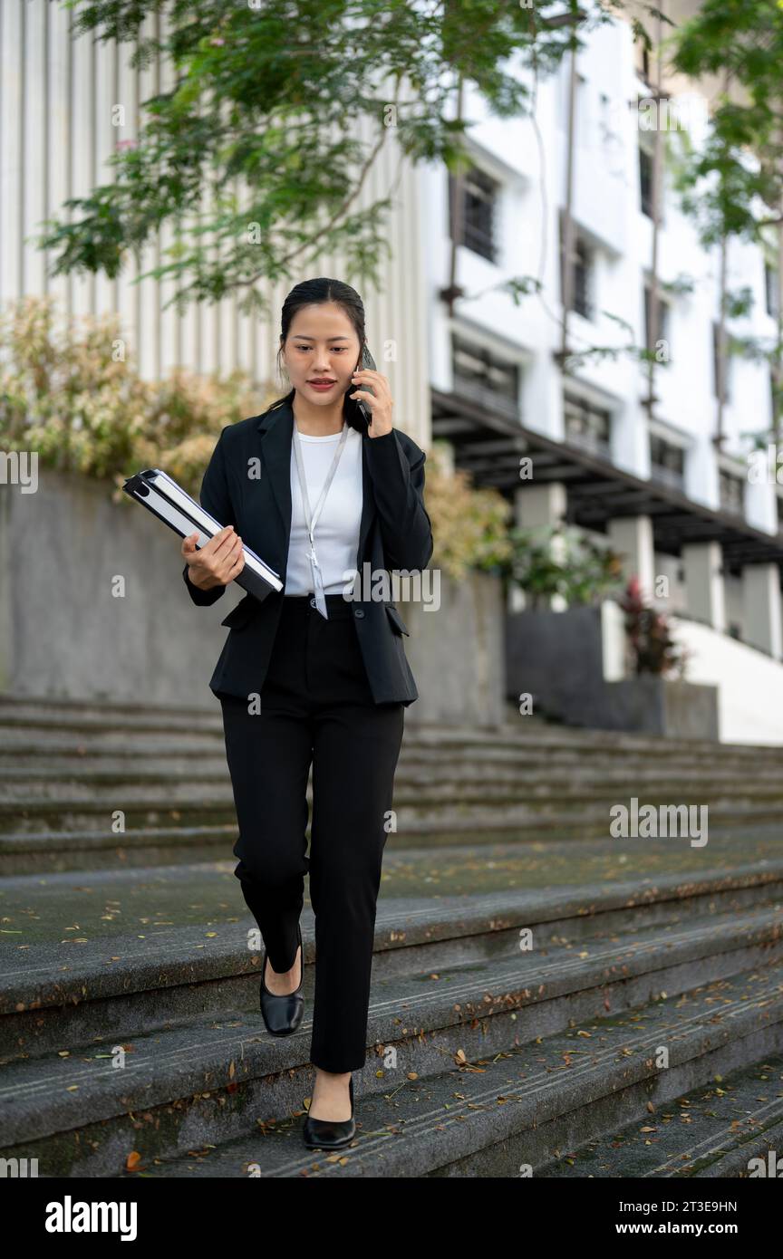 Eine beschäftigte und professionelle Millennials-asiatische Geschäftsfrau in formellem Business-Anzug spricht am Telefon mit einem Kunden, während sie die Treppe i hinuntergeht Stockfoto