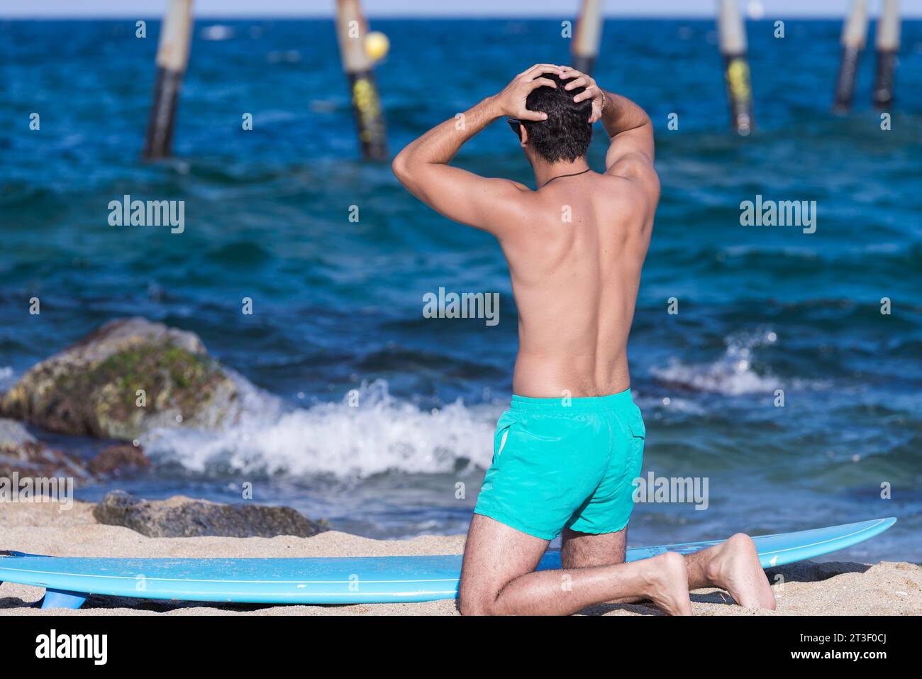 Rückansicht des knienden Mannes am Strand mit seinem Surfbrett, das auf das Meer blickt, bevor er mit den Händen auf dem Kopf surft Stockfoto Rückansicht des knienden Mannes am Strand mit seinem Surfbrett, das auf das Meer blickt, bevor er mit den Händen auf dem Kopf surft Stockfoto