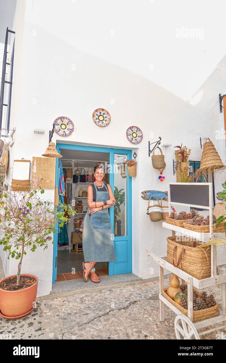 Vorderansicht einer spanischen Besitzerin eines kleinen Souvenirgeschäfts, die stolz vor der Tür ihres Geschäfts posierte. Sie schaut in die Kamera und lächelt. Stockfoto