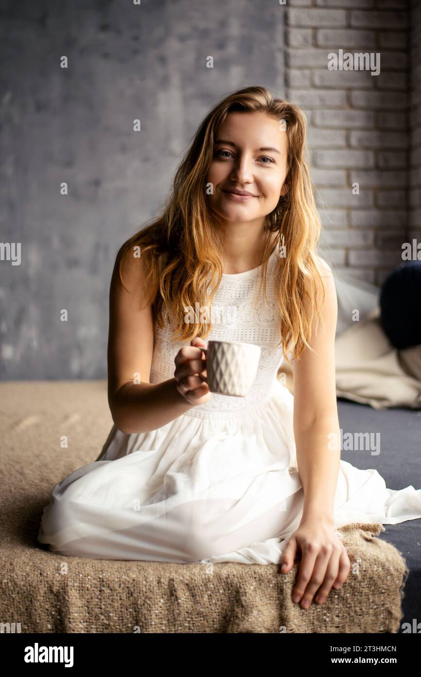 Die junge Frau grinst und hält sich in der Hand eine Kaffeetasse. Das Bild fängt das Wesen des morgendlichen Kaffeererituals ein, strahlt Positivität und Ruhe aus Stockfoto