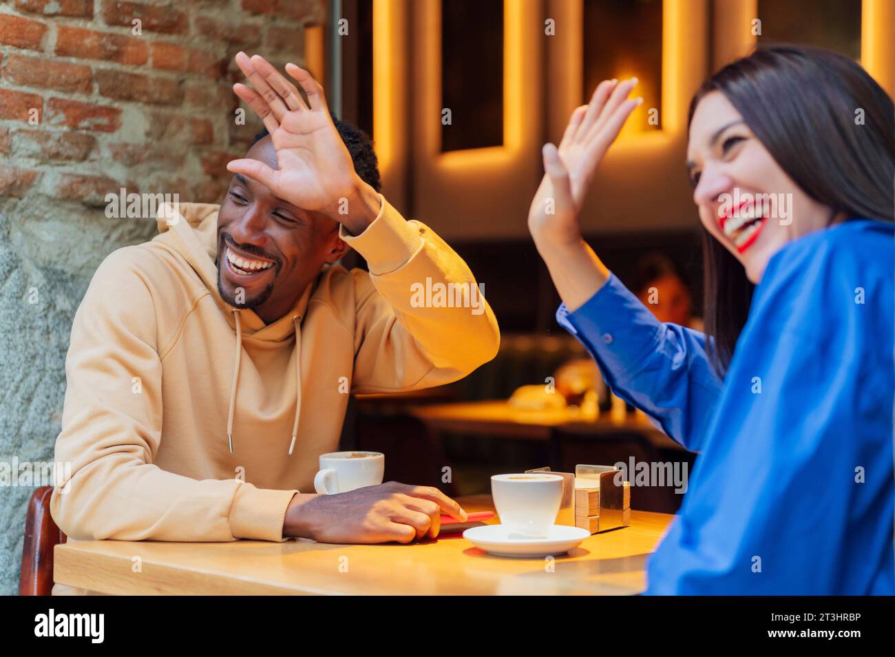 Fröhliches junges Paar mittleren Alters. Afroamerikaner und Kaukasierin beste Freunde, die ihrem neuen Freund winken, während sie sitzen und bre haben Stockfoto