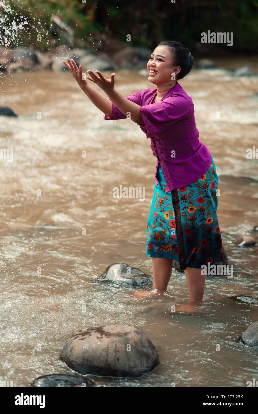 Eine wunderschöne asiatische Frau, die ihre Hand wäscht und mit dem Wasser spielt, während sie in einem traditionellen lila Kleid und grünen Rock in der Nähe des Flusses steht Stockfoto