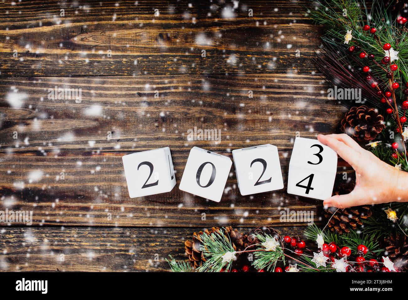 Frauenhand kippt die 2023 Holzkalenderblöcke des Neujahrs auf 2024. Weihnachtsbaumbeleuchtung, Tannenzweige, rote Winterbeeren und Schnee. Draufsicht. Stockfoto