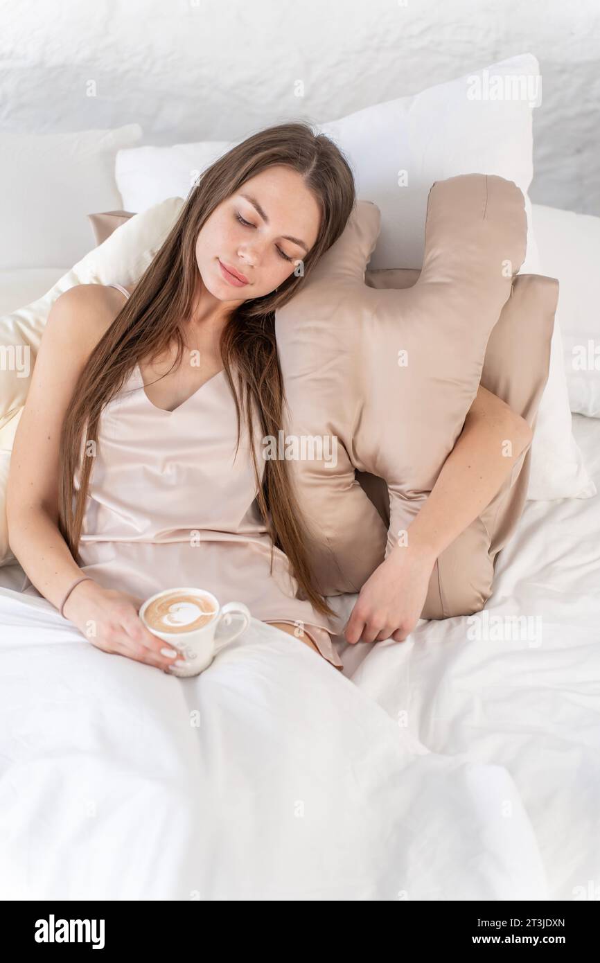 Ein sexy Mädchen mit einem Schönheitskissen und einer Kaffeetasse in den Händen sitzt im Bett. Vertikales Foto Stockfoto