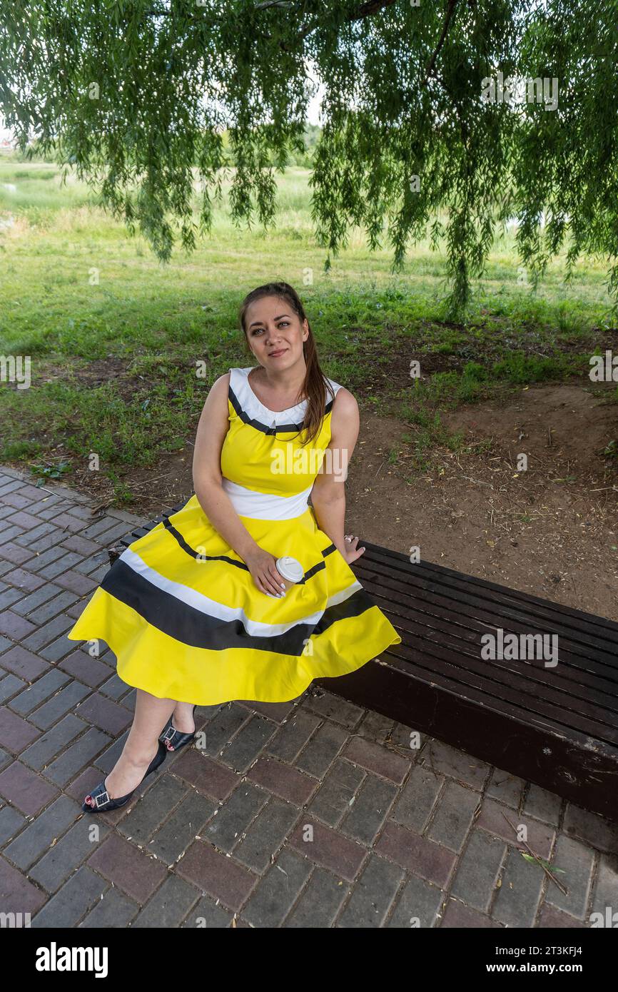 Porträt eines jungen, wunderschönen Mädchens in einem eleganten Kleid mit einer gelben Tasse Kaffee im Park auf einer Bank, eine glückliche und lächelnde Frau Stockfoto