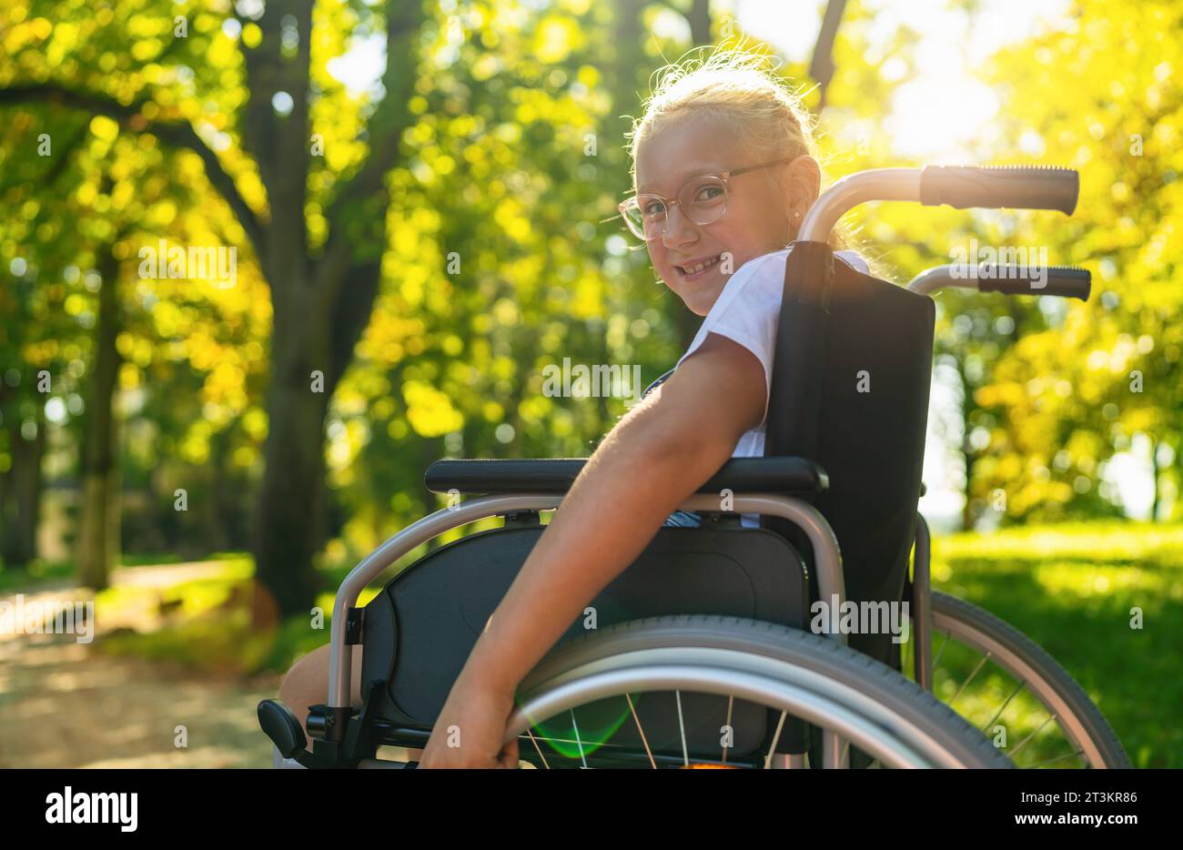 Lächelndes, junges behindertes blondes Mädchen sitzt im Rollstuhl beim Spaziergang im Park, Teenager Behindertenkonzept Bild Stockfoto