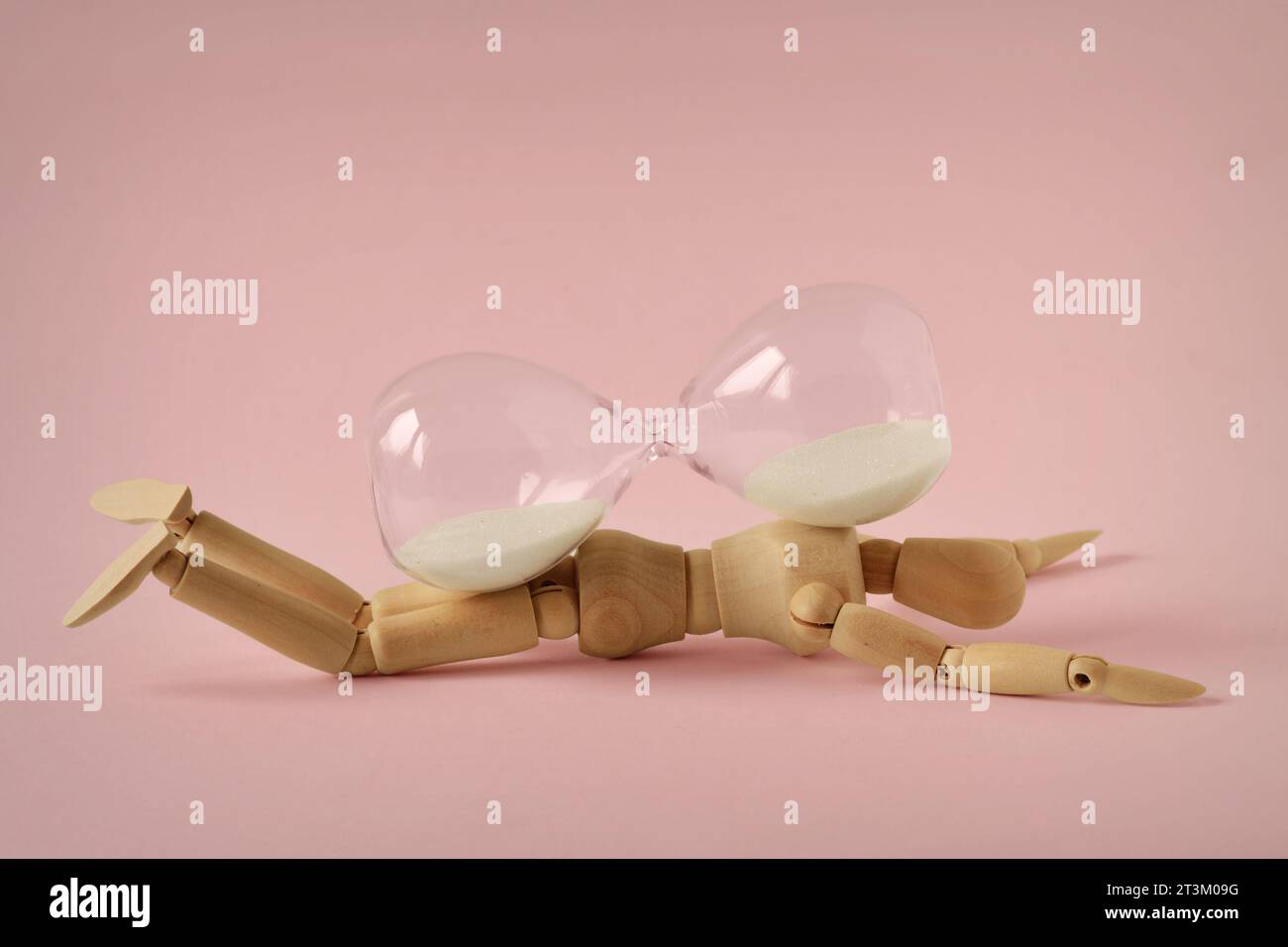 Hölzerne Schaufensterpuppe unter Sanduhr auf rosafarbenem Hintergrund - Konzept der Stoppzeit, Gesundheit und Alterung Stockfoto