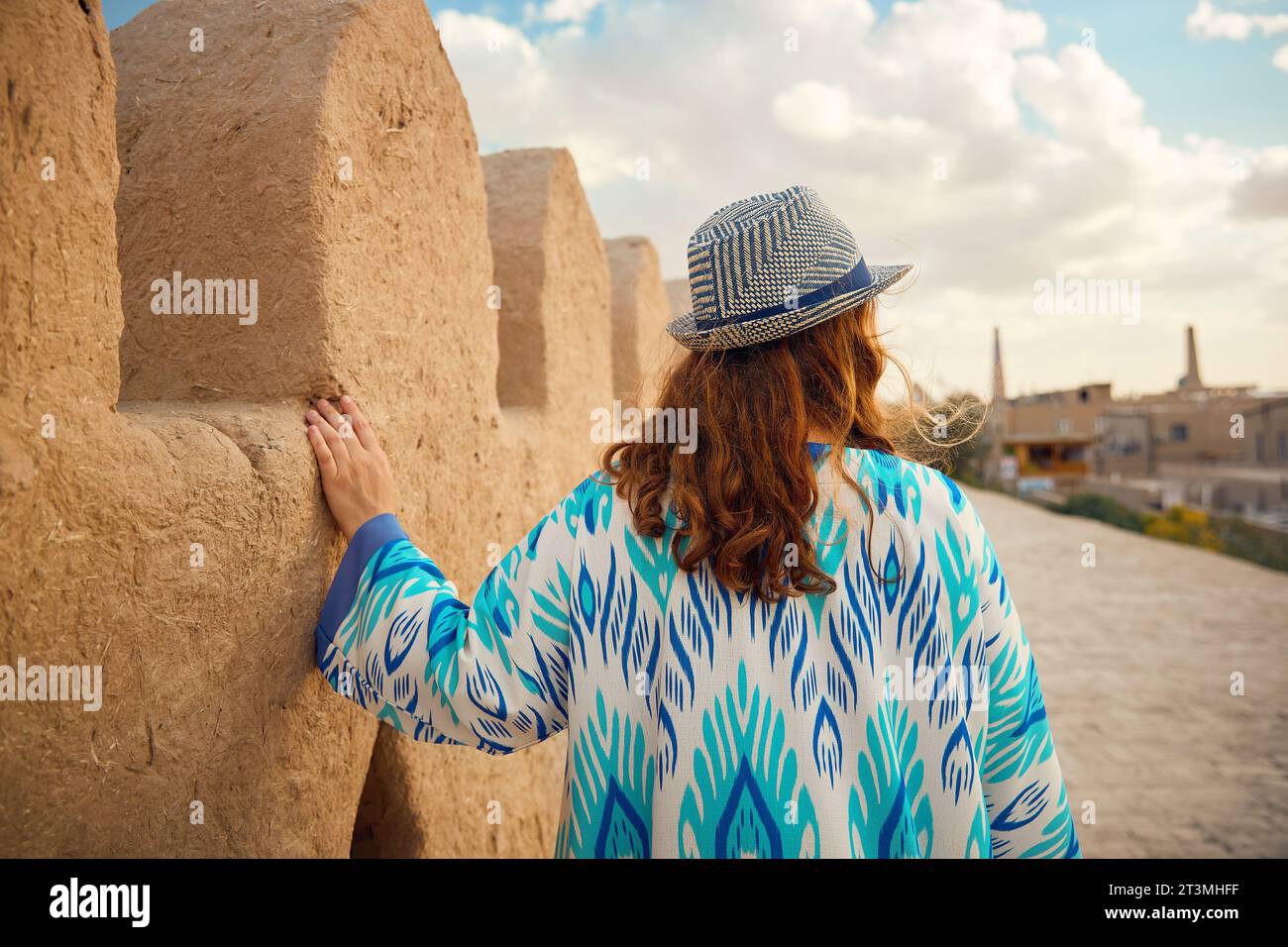 Porträt einer jungen Frau in ethnischer Kleidung mit blauem Ornament und Hut, die an der Stadtmauer in Itchan Kala in der alten Stadt Chiwa in Usbekistan spaziert. Stockfoto