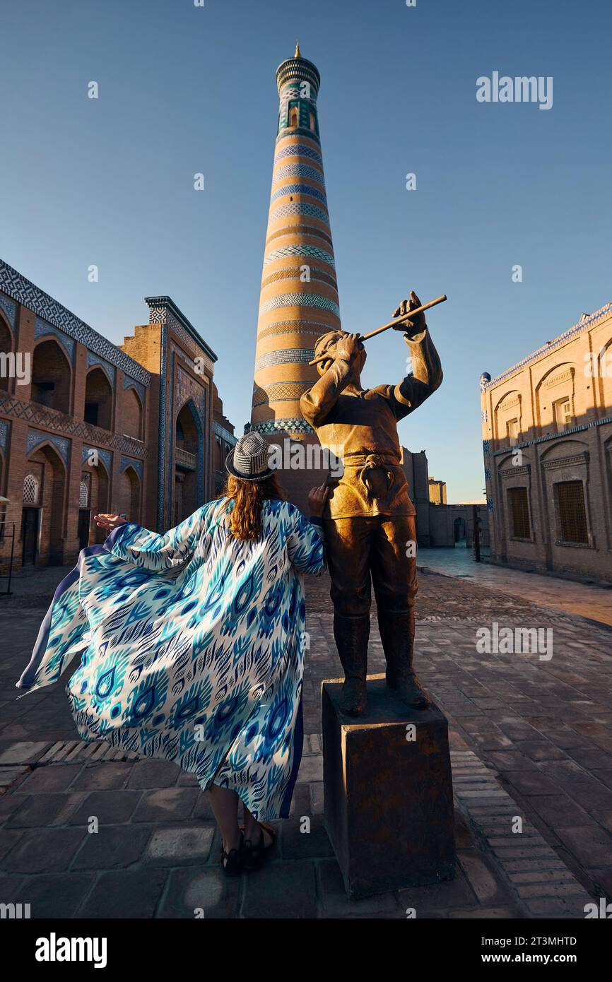 Junge Frau in ethnischer Kleidung mit blauem Ornament und Hut in der Nähe von Flötistenstatue und hohem Minarett auf dem Platz in Itchan Kala alte Stadt Chiwa in Usbekist Stockfoto