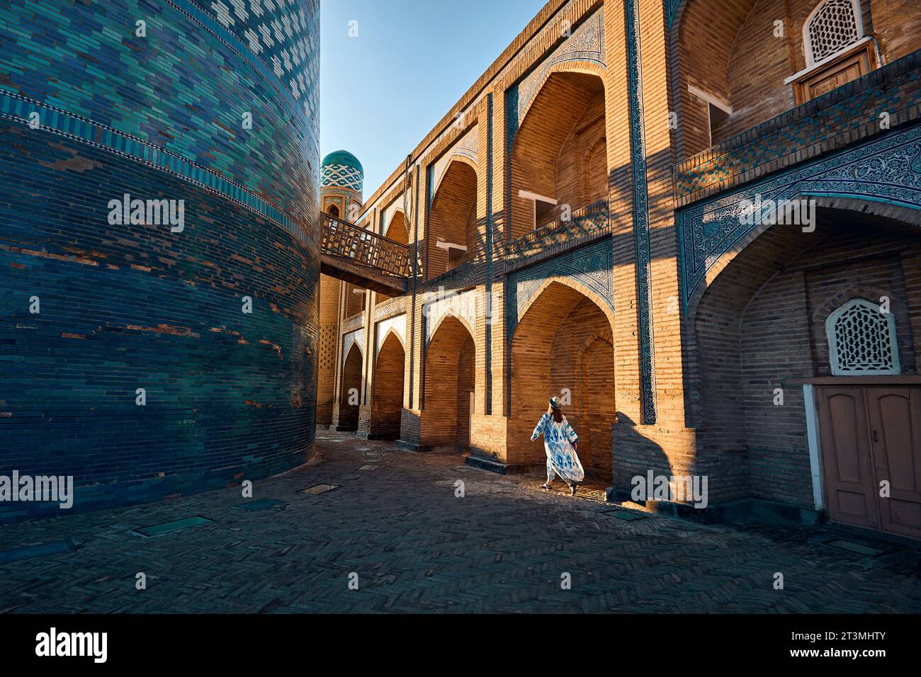 Junge Frau in ethnischer Kleidung mit blauem Ornament und Hut, die in der Nähe des blauen Minaretts Kalta und der alten Madrasah in der alten Stadt Chiwa in Itchan Kala läuft Stockfoto