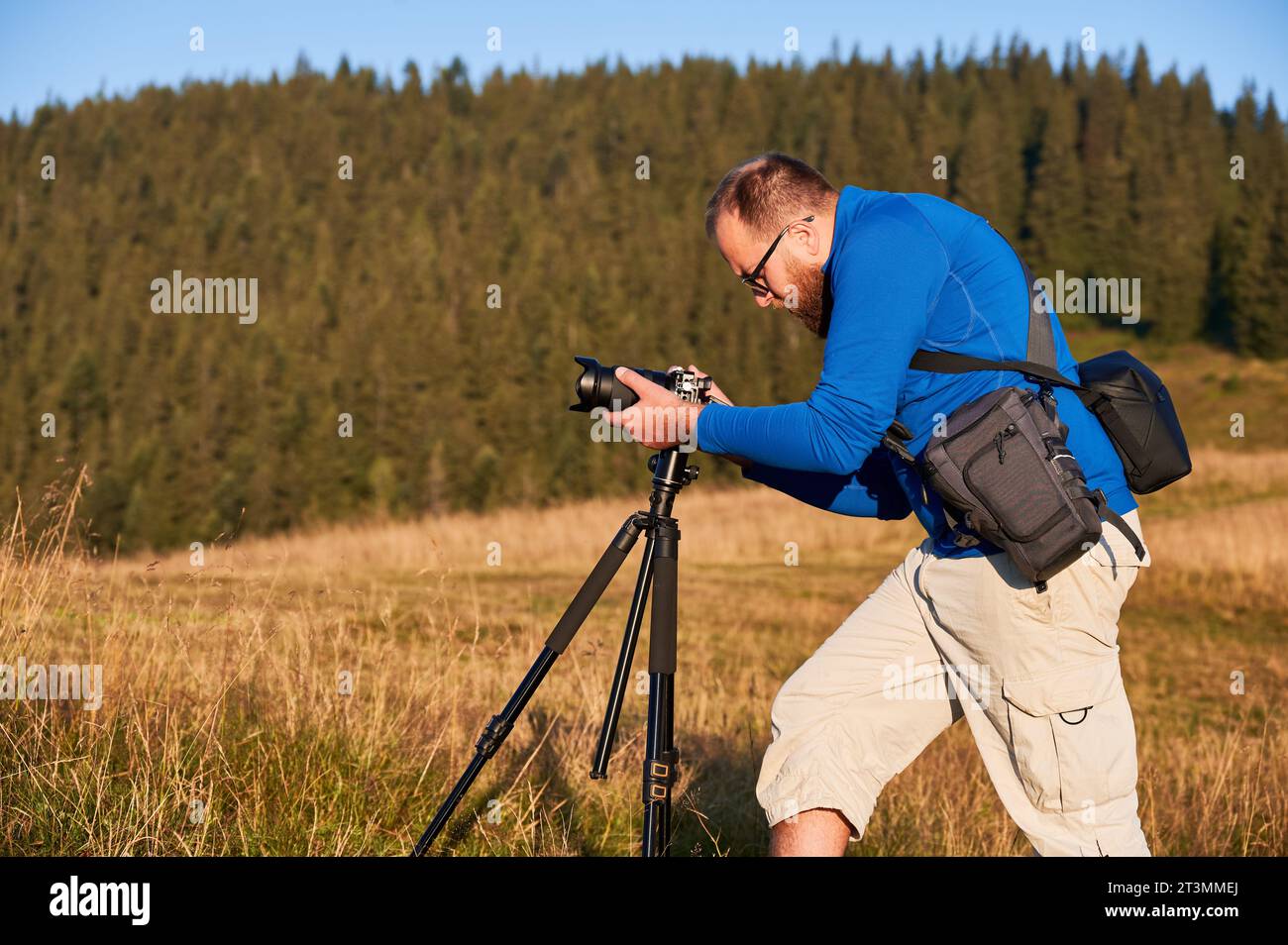 Professioneller Fotograf, der Fotos in den Bergen macht. Mann fokussiert seine Kamera, um ein schönes Foto zu machen. Seitenansicht der jungen männlichen Einstellkamera auf dem Hintergrund von Hügeln. Stockfoto