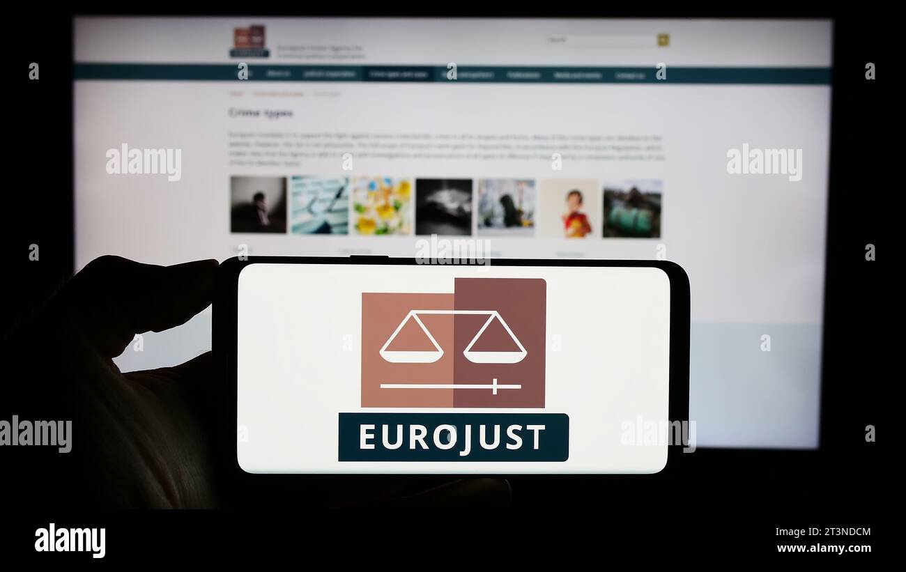 Person, die ein Mobiltelefon mit dem Logo der EU-Agentur für justizielle Zusammenarbeit in Strafsachen (Eurojust) vor der Webseite hält. Schwerpunkt auf dem Display des Telefons. Stockfoto