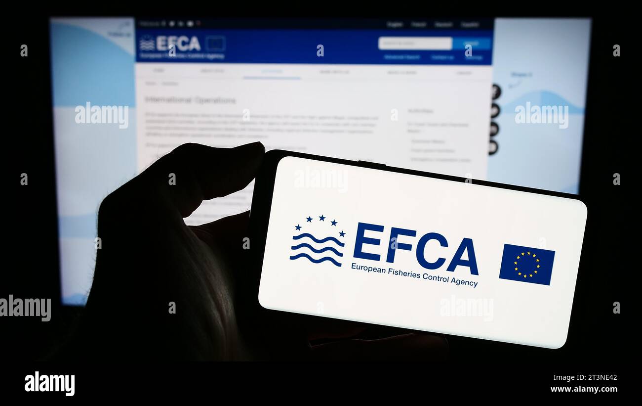 Person mit Mobiltelefon und Logo der Europäischen Fischereiaufsichtsagentur (EFCA) vor der Webseite. Schwerpunkt auf dem Display des Telefons. Stockfoto