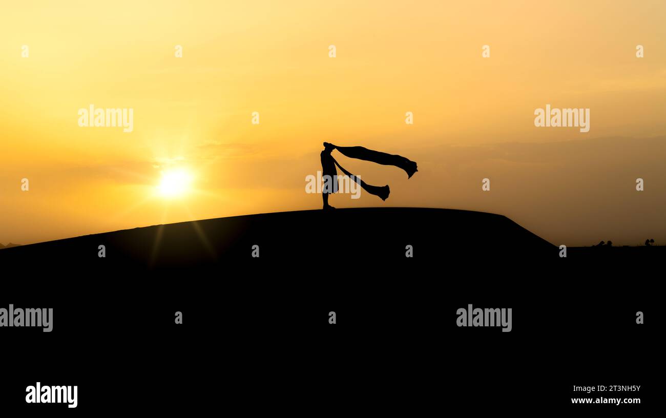 Silhouette eines nicht identifizierten Berbermannes in seinem langen Gewand und Turban, der im Wind auf Sanddünen bei schönem Sonnenuntergang in der Sahara, Marokko, schwingt Stockfoto