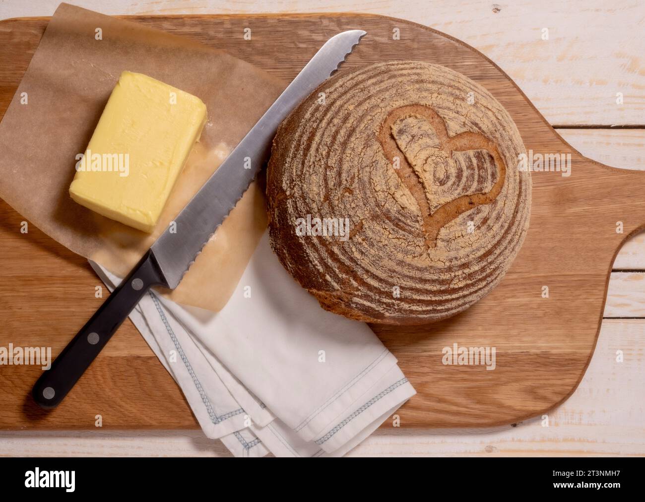 Flache Lage einer glutenfreien Sauerteigboule mit Herzform, die mit einem Butterblock auf eine Holzbrotplatte gelegt wird. Stockfoto