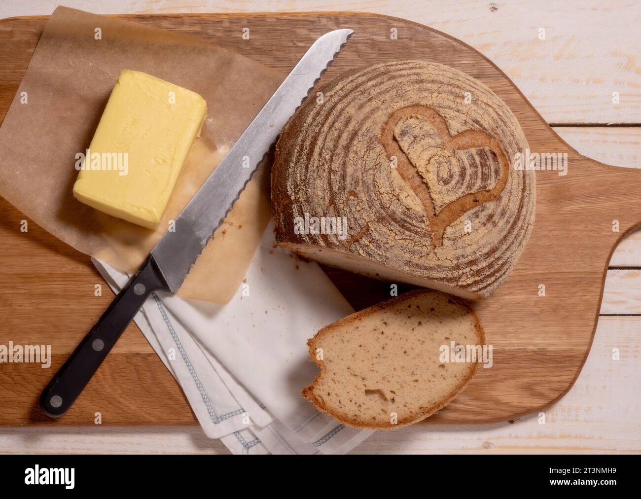 Flache Lage einer glutenfreien Sauerteigboule mit Herzform, die mit einem Butterblock und einem Brotmesser auf ein hölzernes Schneidebrett gelegt wird. Stockfoto