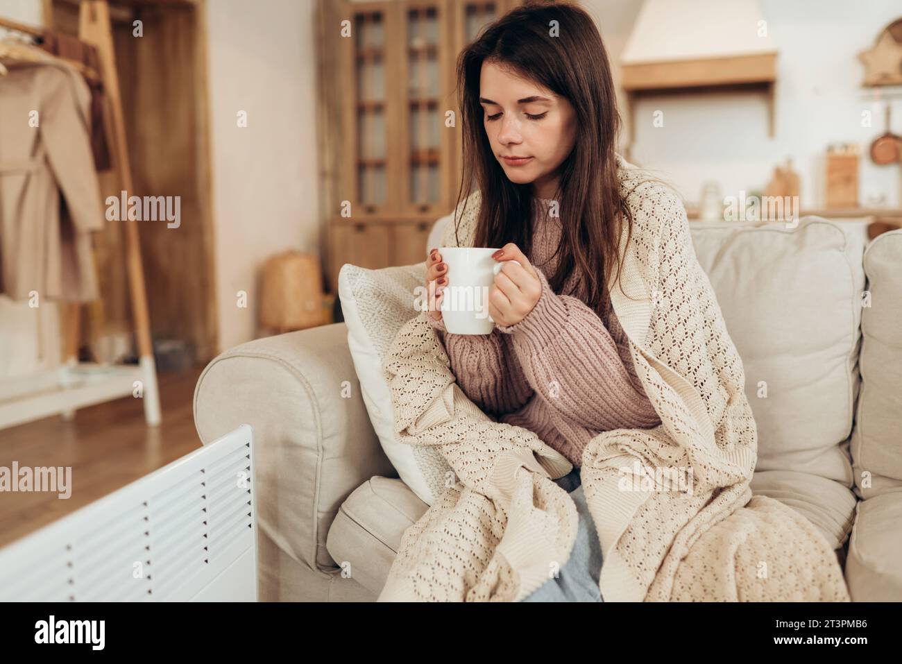 Junge Frau in warmen Kleidern und Karo, die eine Tasse heißes Getränk hält, während sie zu Hause auf der Couch in der Nähe der elektrischen Heizung sitzt. Hochwertige Fotos Stockfoto