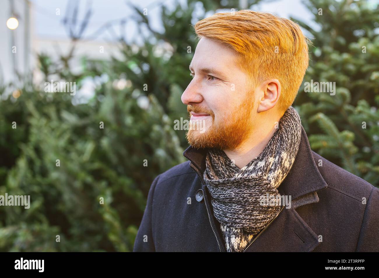 Rothaariger Mann schaut seitlich zwischen weihnachtsbäumen auf einem Straßenmarkt Stockfoto