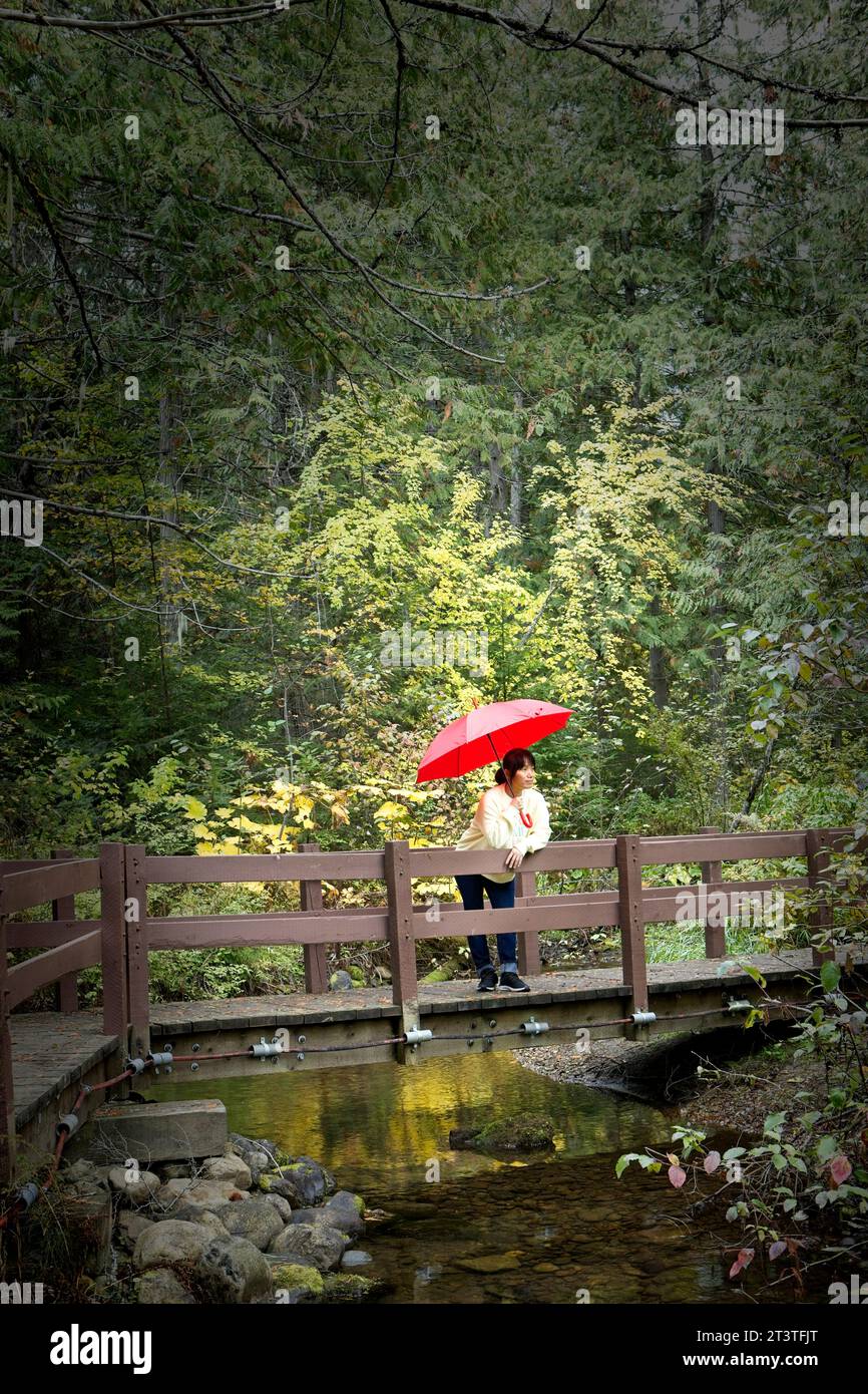 Eine Frau, die einen roten Schirm hält, steht auf einer kleinen Fußgängerbrücke, die über einen Fluss in der Nähe des Robinson Lake im Norden von Idaho überspannt. Stockfoto